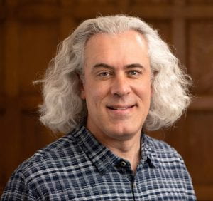 Headshot of Rogers, with curly grey hair approaching shoulder length and a blur plaid shirt. Wooden panel background. © Photo by Mara Lavitt