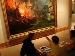 woman and children sitting on floor looking up at large oil painting of ships of fire