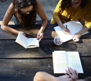 Three people sitting at a wooden picnic table reading books and taking notes.