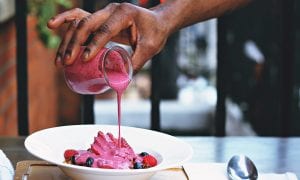 An image of a hand pouring a pink-colored smoothie onto a bowl of fresh fruit.